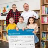 Fünf Personen stehen vor einem Bücherregal und halten einen großen Spendenscheck in der Hand. Sie haben eine Bücherei neu eröffnet.