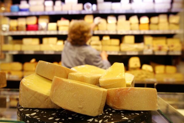 Käse ist auf der Käsetheke in einem Supermarkt zu sehen, während im Hintergrund eine Mitarbeiterin Käse aus einem Regal nimmt.