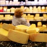 Käse ist auf der Käsetheke in einem Supermarkt zu sehen, während im Hintergrund eine Mitarbeiterin Käse aus einem Regal nimmt.