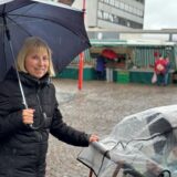 Eine Frau mit Regenschirm, lächelt. Sie schiebt einen Kinderwagen vor sich. Im Hintergrund ist ein Marktstand zu sehen.