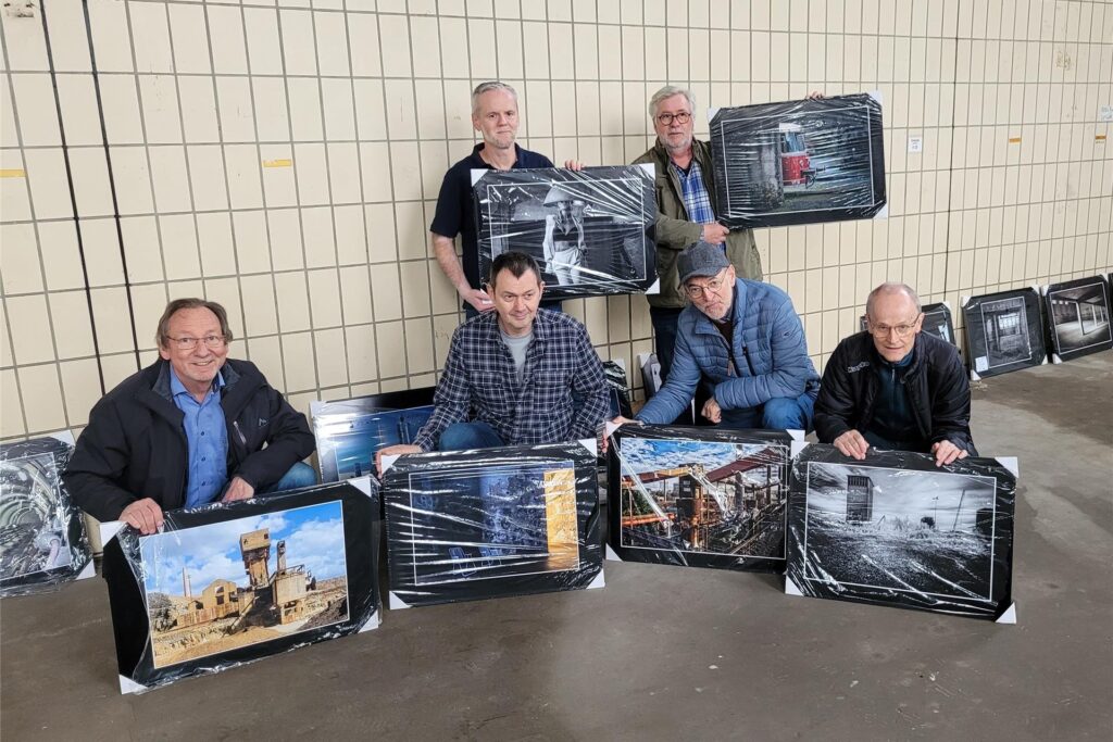 In einer Halle hocken vier Männer, die jeweils ein Foto vor sich auf dem Boden gestellt haben. Hinter ihnen stehen zwei Männer, die auch jeweils einen Bilderrahmen in der Hand halten. In einer Halle hocken vier Männer, die jeweils ein Foto vor sich auf dem Boden gestellt haben. Hinter ihnen stehen zwei Männer, die auch jeweils einen Bilderrahmen in der Hand halten.