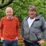 Zwei Männer stehen in einem Garten vor einem Baum. Hinter ihnen sind Sitzmöbel aus Holz und neben ihnen ist ein Tisch zu sehen.