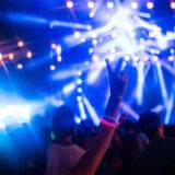 Im Vordergrund ist ein Arm mit knallig bunten Armbändern in die Luft gestreckt. Zeige und Mittelfinger, zeigen in die höhe. Im Hintergrund sind viele Mneschen zu erkennen und blaue und weiße strahlende Lichter.