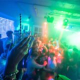 Eine Hand im Vordergrund hält ein Getränk mit Strohhalm hoch. Im Hintergrund die Szene einer Party. Viele Menschen auf einer Tanzfläche, bunte Lichter.