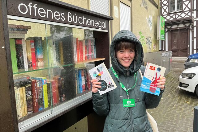 RZ-Autorin Janka Hardenacke hält zwei Bücher in der Hand und steht vor dem offenen Bücherregal vor dem Gasthaus in Recklinghausen.
