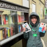 RZ-Autorin Janka Hardenacke hält zwei Bücher in der Hand und steht vor dem offenen Bücherregal vor dem Gasthaus in Recklinghausen.