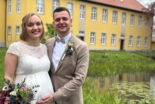 Braut und Bräutigam stehen dicht nebeneinander. Sie trägt weiß, er beige. In ihrer rechten Hand hält die Braut einen Blumenstrauß. Er hat seine rechte Hand auf ihren Babybauch gelegt.