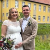 Braut und Bräutigam stehen dicht nebeneinander. Sie trägt weiß, er beige. In ihrer rechten Hand hält die Braut einen Blumenstrauß. Er hat seine rechte Hand auf ihren Babybauch gelegt.