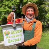 Frank Hoffmann halt sich bunt verkleidet und hält einne Eimer mit dem Schriftzug Gustav, der schnelle Gärtner in seinen Händen.