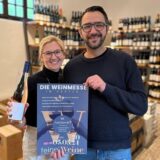 Sandra Leise und Sebastian Banhold lächeln in die Kamera. Sie hält eine Flasche Wein, er ein Plakat der Wein-Messe. Im Hintergrund steht ein Weinregal.