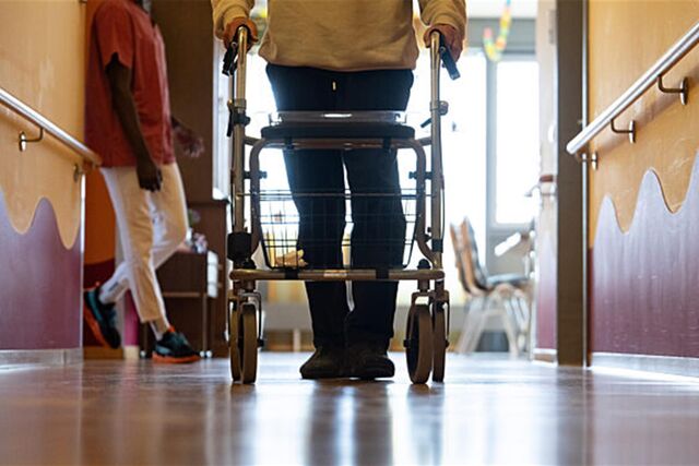 Eine ältere Person läuft mit einem Rollator über den Flur in einem Pflegeheim. Im Hintergrund steht eine Pflegerin.