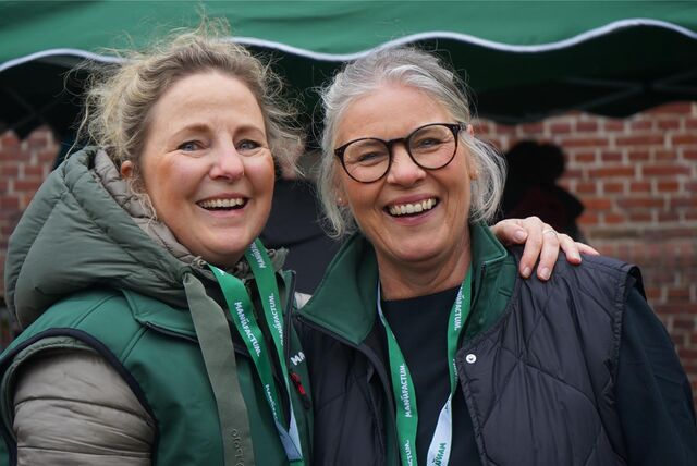 Zwei Frauen stehen nebeneinander und blicken lachend in die Kamera. Im Hintergrund ist ein Pavillon und ein verklinkertes Gebäude zu erkennen.