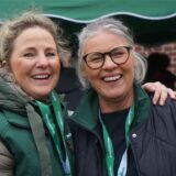 Zwei Frauen stehen nebeneinander und blicken lachend in die Kamera. Im Hintergrund ist ein Pavillon und ein verklinkertes Gebäude zu erkennen.