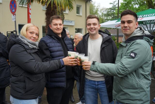 Drei Männer und eine Frau stehen nebeneinander, prosten sich mit Bier in Plastikbechern zu und blicken in die Kamera.