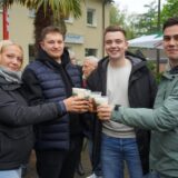 Drei Männer und eine Frau stehen nebeneinander, prosten sich mit Bier in Plastikbechern zu und blicken in die Kamera.