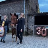 Zwei Frauen stehen neben einem fuchsfarbenen Pony. Die linke hält einen Champanger förmigen Ballon in der Hand. Im Hintergrund sind Pferdeboxen zu erkennen. Eine ist mit einer großen Dreißig geschmückt.