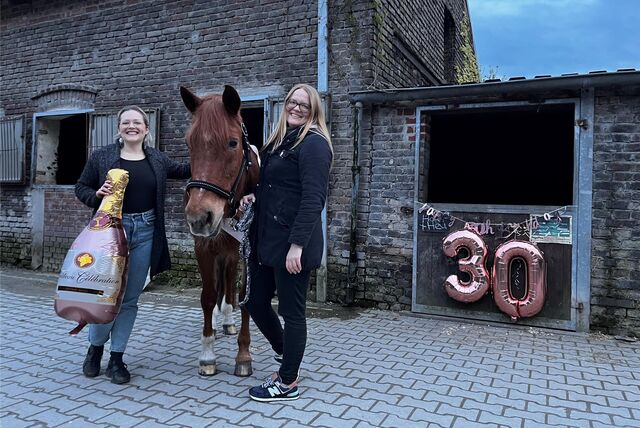 Zwei Frauen stehen neben einem fuchsfarbenen Pony. Die linke hält einen Champanger förmigen Ballon in der Hand. Im Hintergrund sind Pferdeboxen zu erkennen. Eine ist mit einer großen Dreißig geschmückt.