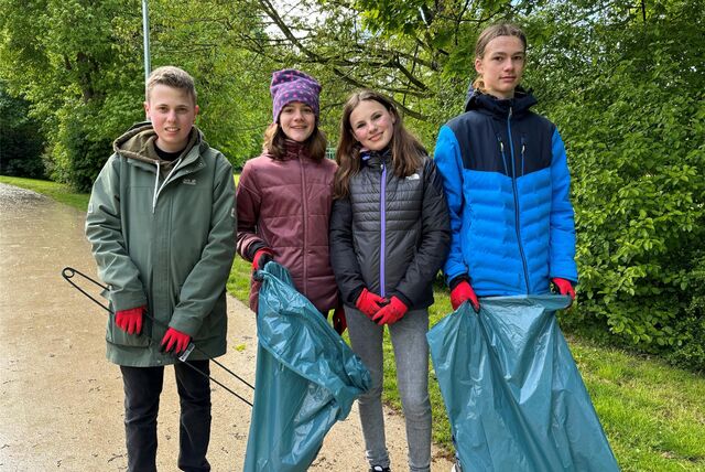 Zwei Jungen und zwei Mädchen stehen vor grüner Kulisse. Sie sind mit roten Handschuhen, blauen Müllsäcken und Greifzangen ausgestattet.