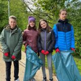 Zwei Jungen und zwei Mädchen stehen vor grüner Kulisse. Sie sind mit roten Handschuhen, blauen Müllsäcken und Greifzangen ausgestattet.