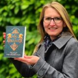 Britt Glaser präsentiert „Das Herz von Arkamoor“. Sie hält das Buch vor einem grünen Hintergrund in ihren Händen.