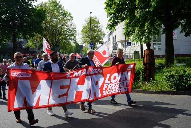 Menschen demonstrieren und tragen ein rotes Banner mit der Forderung „Mehr Lohn, mehr Freizeit, mehr Sicherheit“.