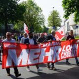 Menschen demonstrieren und tragen ein rotes Banner mit der Forderung „Mehr Lohn, mehr Freizeit, mehr Sicherheit“.