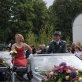 Ein festlich gekleidetes Paar sitz in einem geschmückten Cabriolet. Die Frau trägt ein rotes Kleid und einen Blumenstrauß, der Mann eine Schützenuniform.