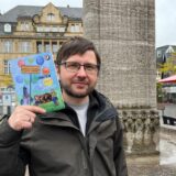 Kamill Eliasch steht vor dem Reiterbrunnen in Castrop-Rauxel und zeigt sein Kinderbuch „Caratup Raukel mit Tomatensaft“.