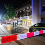 Rot-weißes Polizeiabsperrband vor der in der Nacht hell beleuchteten Bankfiliale, deren Frontscheiben zerstört sind.