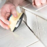 Ein Mann reinigt mit einem gelben Schlamm die Fugen im Badezimmer – hier hat sich Schimmel gebildet.