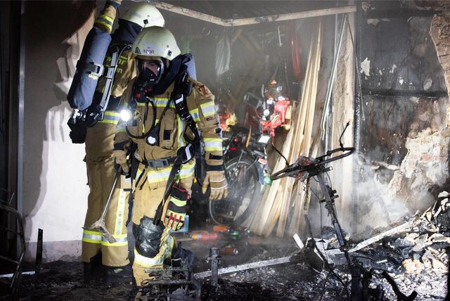 Feuerwehrleute stehen vor einer Garage und dem verkohlten Wrack eines E-Scooters.