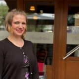 Eine Frau mit kurzen blonden Haaren lächelt. Im Hintergrund sind Fenster eines Lokals zu erkennenn.
