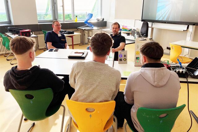 Benjamin Litzenberger (rechts) und Michael Dertmann der Firma Liftec aus Marl sitzen in einem Klassenzimmer der Hertener Willy-Brandt-Schule an einem Tisch.  Vor ihnen sind drei Jugendliche, mit denen sie sich unterhalten.