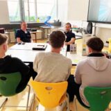Benjamin Litzenberger (rechts) und Michael Dertmann der Firma Liftec aus Marl sitzen in einem Klassenzimmer der Hertener Willy-Brandt-Schule an einem Tisch.  Vor ihnen sind drei Jugendliche, mit denen sie sich unterhalten.