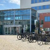 Links sind ein Rathaus und Fahrradbügel zu sehen, reichte ein Mann mit Brille, der in die Kamera blickt. (Montage)