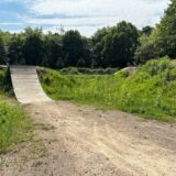 Eine Strecke, die zu einer steilen Holzrampe führt, ist zu sehen. Der Weg liegt mitten im Grünen. Im Hintergrund stehen Bäume.