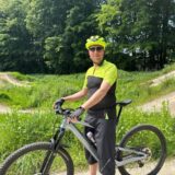 Ein Mann auf einem Fahrrad. Er trägt einen neongrünen Helm und Sportkleidung. Im Hintergrund sind Hügel und Radstrecken zwischen Grünflächen zu sehen.