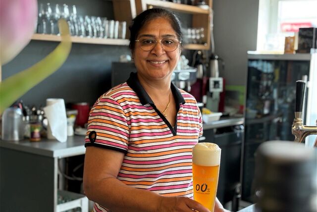Eine Frau im gestreiften T-shirt hält ein Bier in der Hand. Im Hintergrund ist eine Küchenzeile und ein Regal mit Gläsern erkennbar.