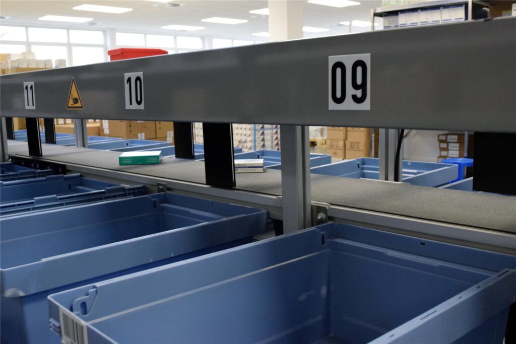 Fließband mit Medikamenten-Schachteln zwischen blauen Kunststoffkisten für die Krankenhaus-Stationen.