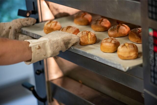 Ein Bäcker holt in der Backstube einer Bäckerei ein Blech mit Brötchen aus dem Ofen.