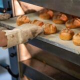 Ein Bäcker holt in der Backstube einer Bäckerei ein Blech mit Brötchen aus dem Ofen.