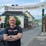 Ein Mann steht an einer Straße, über ihm hängt ein Schild für ein Schützenfest.