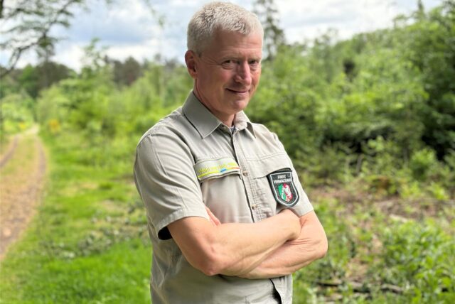 Der Förster steht in seiner Uniform vor einem Waldweg. Die Fläche im Hintergrund ist mit kleinen Bäumen bepflanzt.