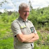 Der Förster steht in seiner Uniform vor einem Waldweg. Die Fläche im Hintergrund ist mit kleinen Bäumen bepflanzt.