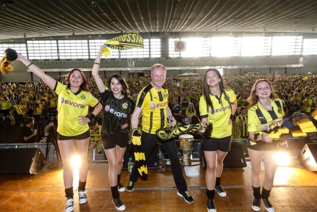 Vier Frauen und ein Mann in BVB-Trikots und mit BVB-Schals tanzen auf einer Bühne.