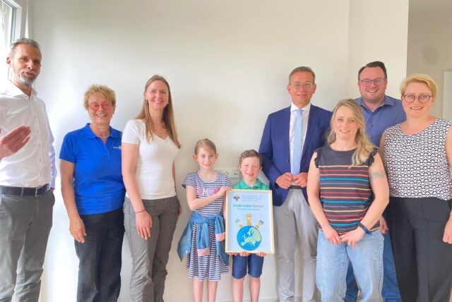 Sieben Erwachsene und zwei Kinder stehen nebeneinander vor einer weißen Wand. DIe Kinder halten ein Bild mit der Zeichnung einer Weltkugel hoch.