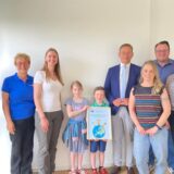 Sieben Erwachsene und zwei Kinder stehen nebeneinander vor einer weißen Wand. DIe Kinder halten ein Bild mit der Zeichnung einer Weltkugel hoch.