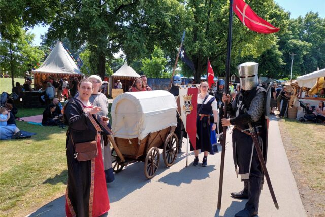 Männer und Frauen in mittelalterlich anmutenden Kostümen ziehen durch einen Park.