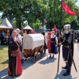 Männer und Frauen in mittelalterlich anmutenden Kostümen ziehen durch einen Park.