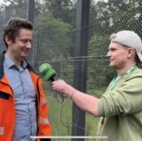 Ein Mann mit einer orangenen Warnweste und ein Reporter mit Mikrofon stehen vor einem Zaun.